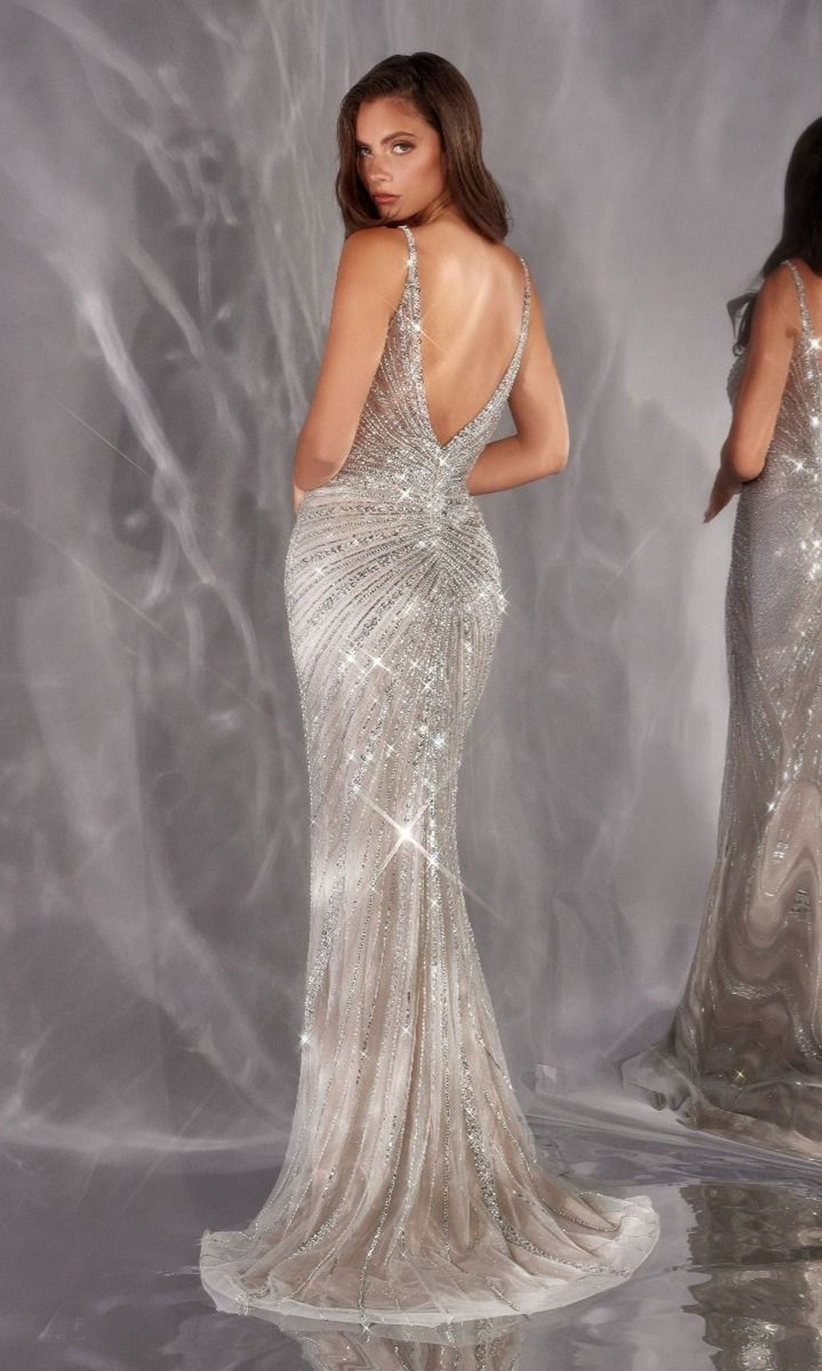 Wearing the Ladivine CC498 long prom dress, a woman in a silver, sequined beaded gown with a deep V-back and fit-and-flare silhouette stands facing away from the camera in front of a reflective backdrop.